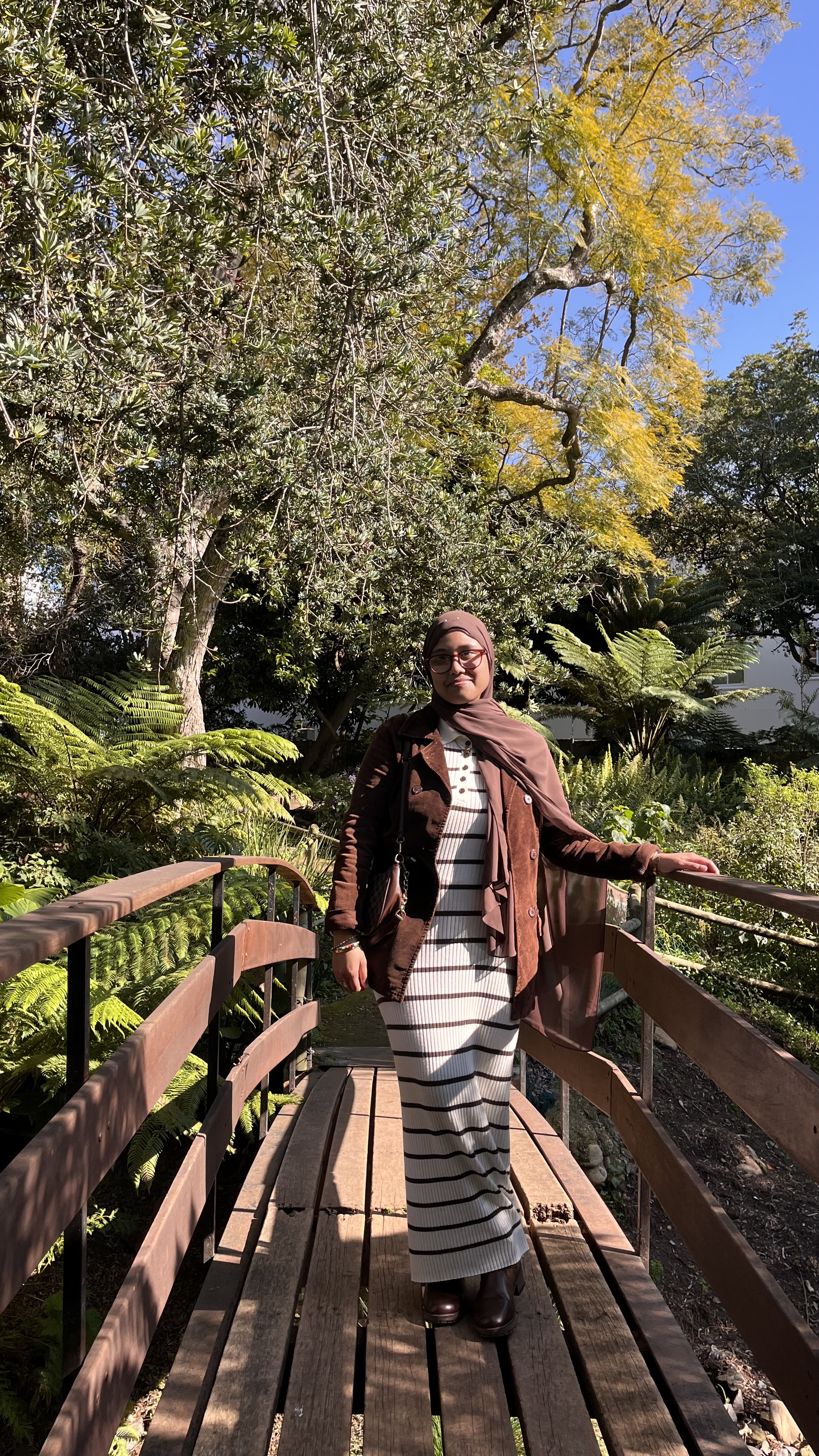 Haanim Pietersen standing on a wooden garden bridge in a brown hijab, suede jacket, and striped dress